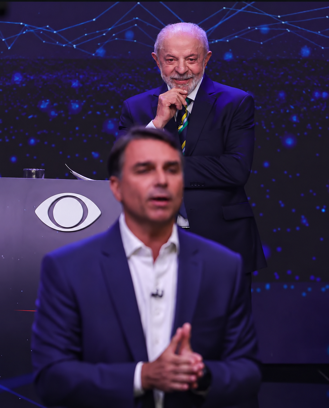 Lula ao fundo e Flávio Bolsonaro em primeiro plano durante debate presidencial